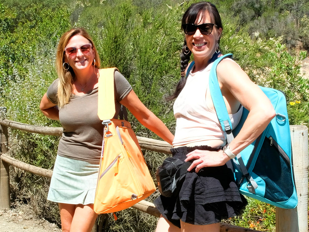 Two woman stop to smile for the camera while they carry their Playamigo ground chairs on a hike outdoors.