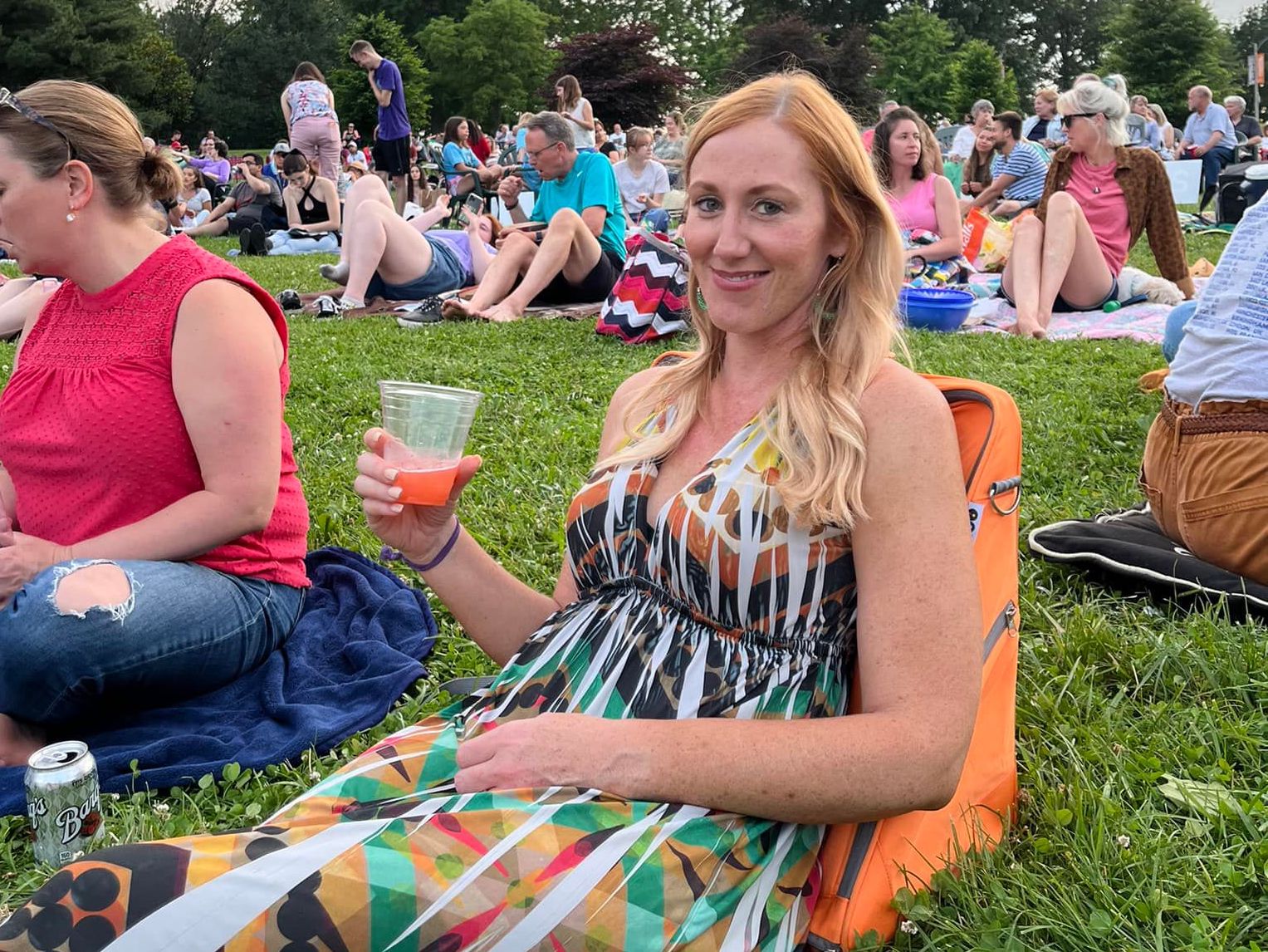 A woman enjoys a drink using her Playamigo ground chair on the grass for Shakespeare in the Park.